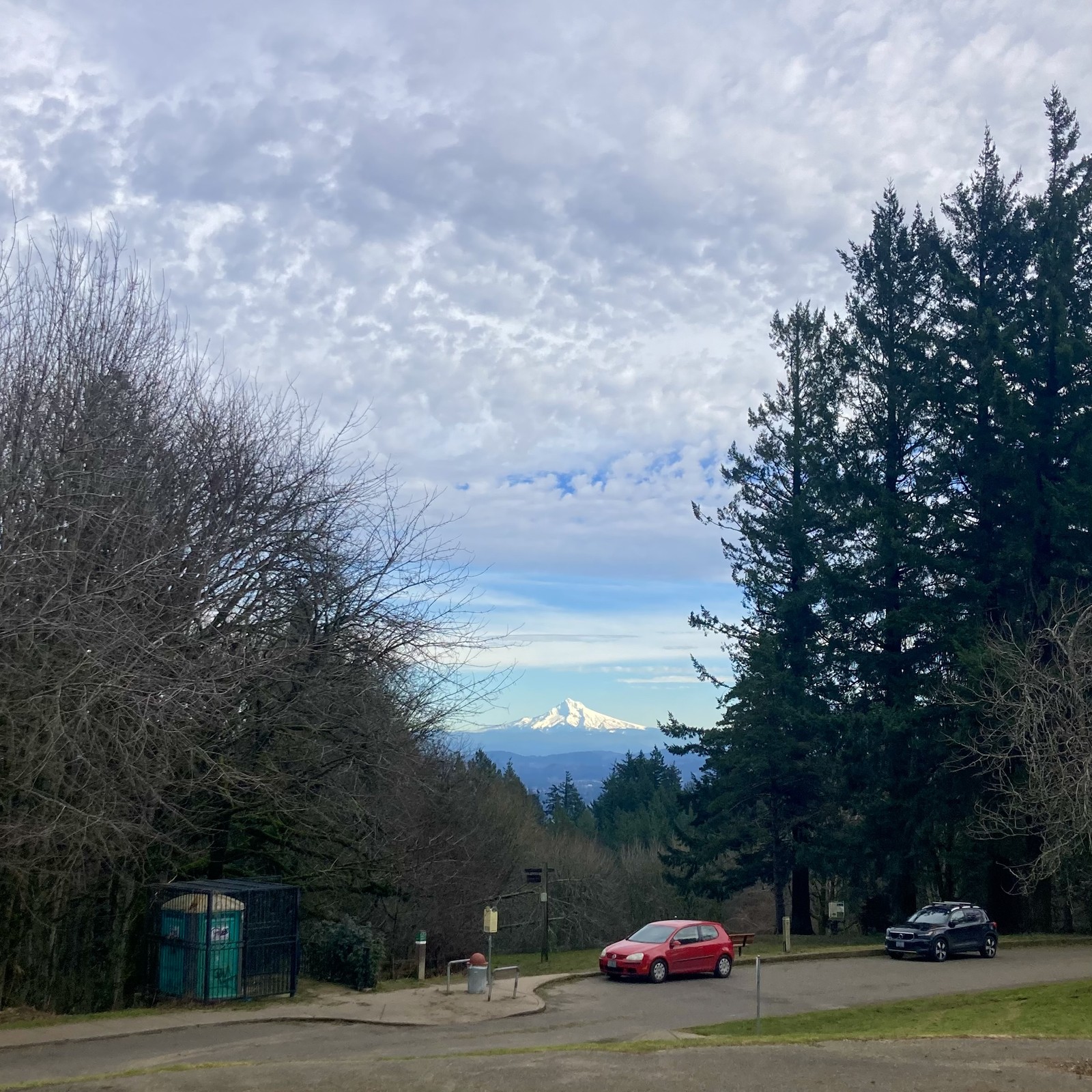On the horizon, Mt. Hood glows with fresh snow under a blue sky. Overhead a layer of bumpy stratus clouds marches slowly northeastward. In the near foreground is a bright red subcompact and a portapotty