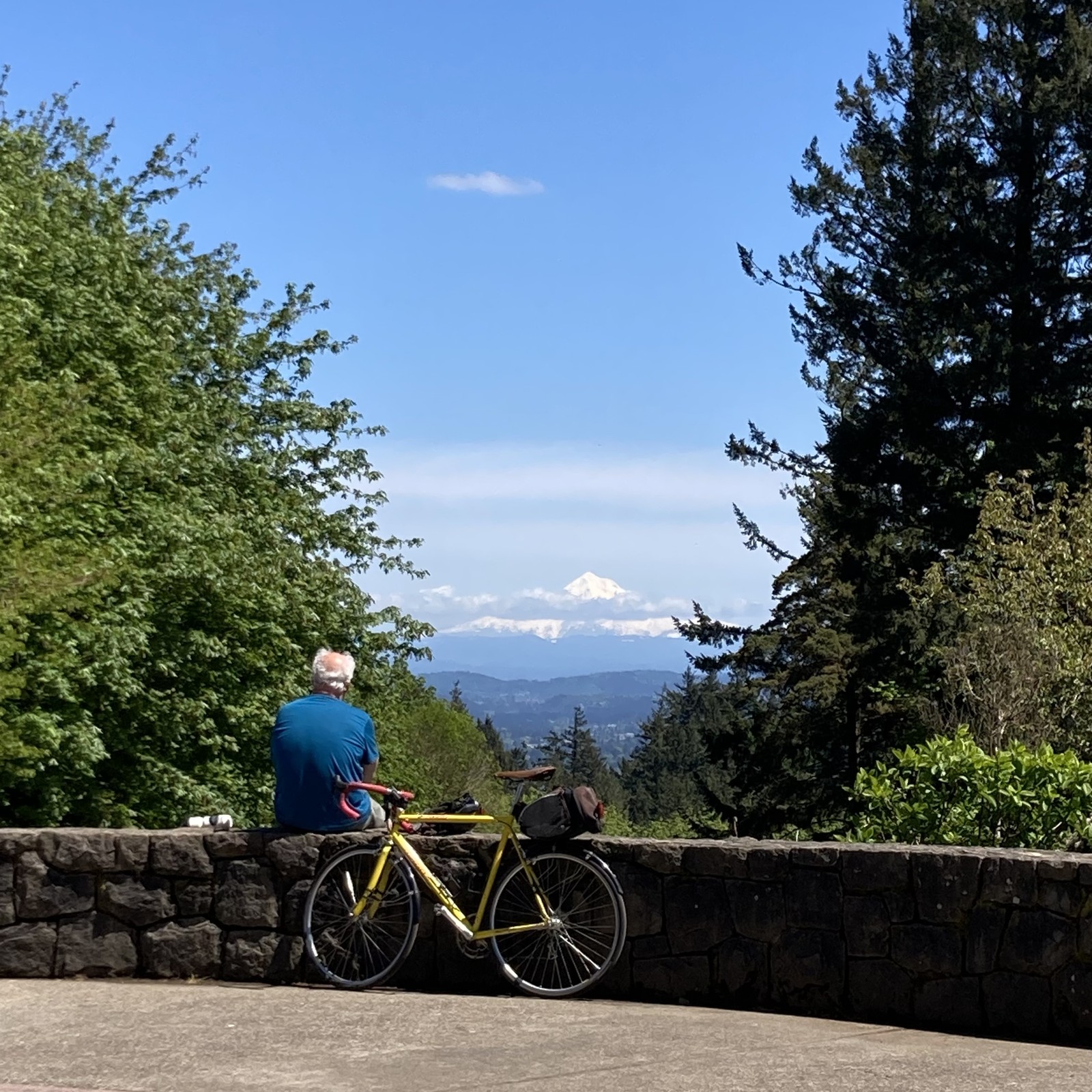 A middle aged man in a t shirt sits with his back to the camera next to a vintage bicycle. He is looking toward mt Hood, whose brilliant white peak is ringed with one low cloud. The sky is otherwise deep and clear