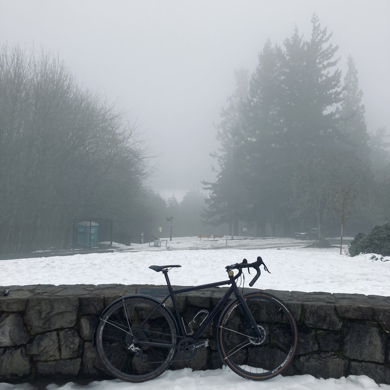 View from Council Crest Park in the direction of Mt. Hood, which is not visible. Week-old icy snow, well-churned by thousands of footsteps, blankets the ground. A cold heavy mist hangs in the air — the inside of a dark cloud clinging to the hilltop. In the foreground, a bicycle leans against a low decorative stone wall. A stand of tall Douglas fir trees about 100' away looms in the mist.