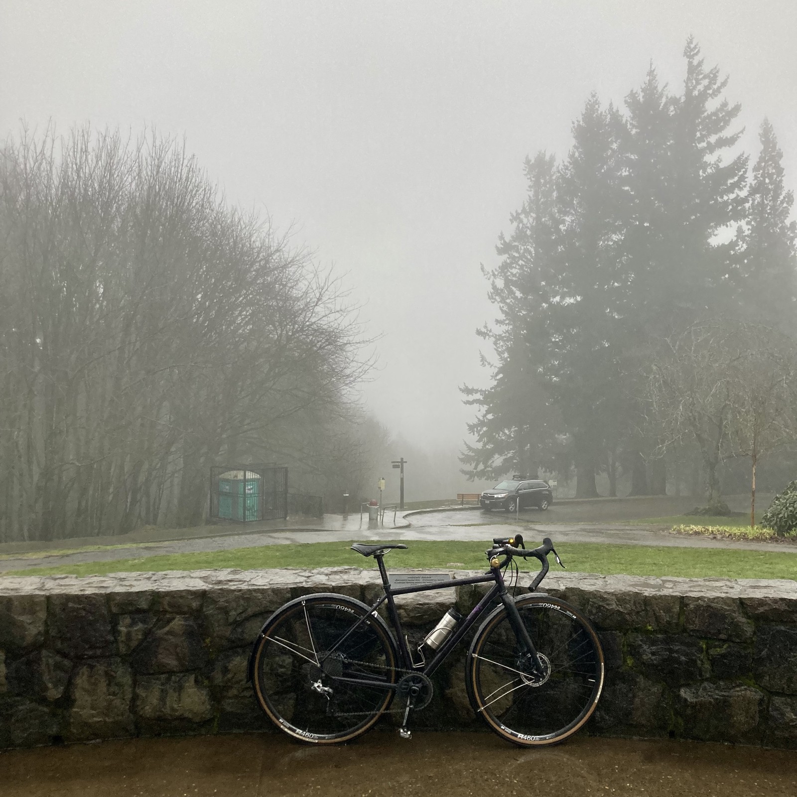View from Council Crest Park toward Mt. Hood which is obscured behind an “atmospheric river” i.e heavy rain from the Pacific. A bicycle leans against a low decorative stone wall in the foreground. A single SUV is parked about 50' away and somewhat downhill. The ground is very wet, the sidewalks have become small brooks of rain.