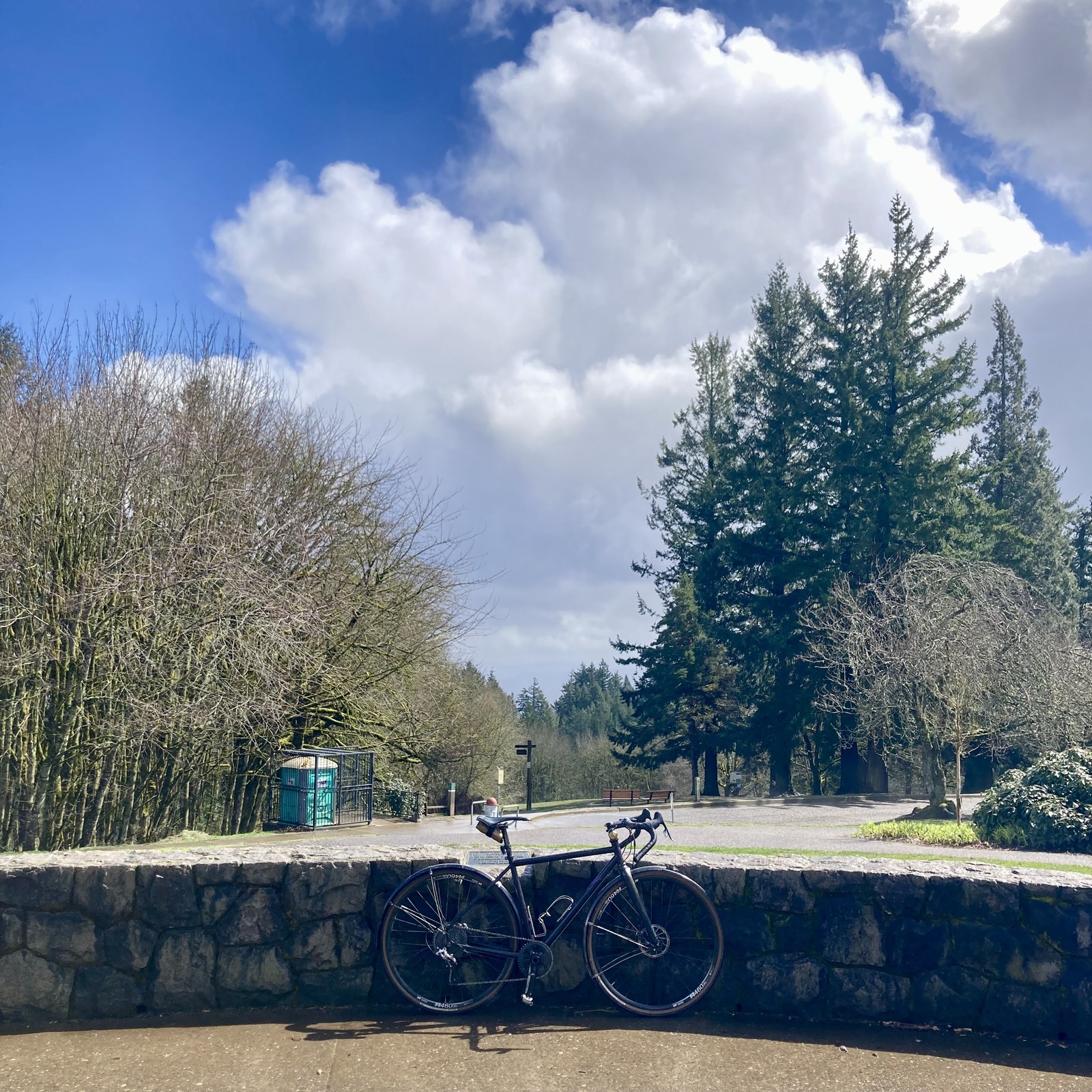 View from Council Crest Park toward Mt. Hood, which is hidden behind many clouds. In the foreground a bicycle leans against a low stone wall. Clouds heavy with rain shine brightly in the early afternoon sunlight. It is a short sunbreak between heavy thunderstorms.