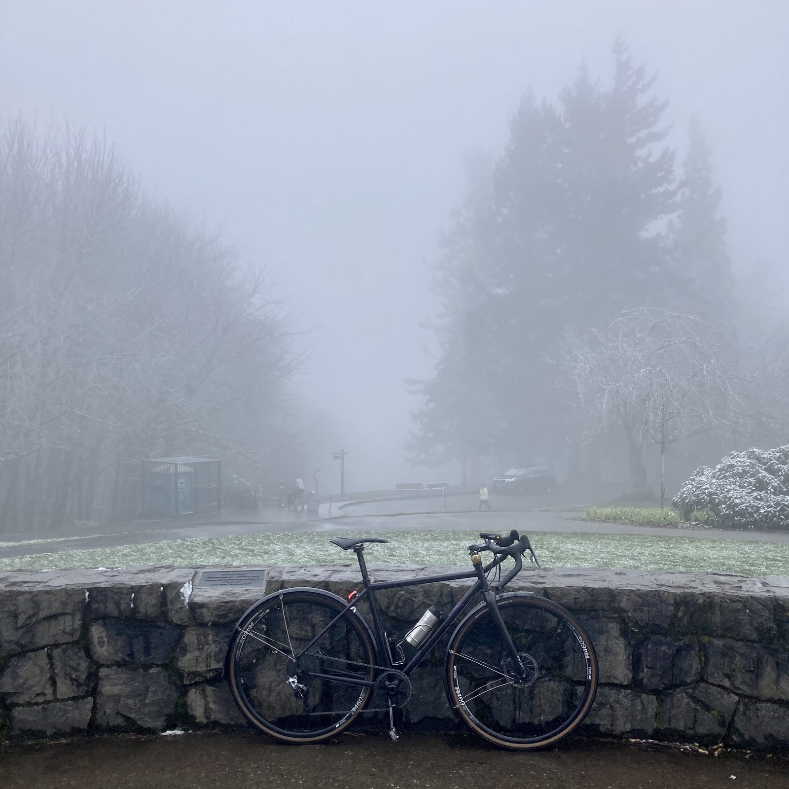 View from Council Crest Park toward Mt. Hood, which is not visible behind a heavy fog that I can only describe as “stationary snow.” In the foreground, a bicycle leans against a low decorative stone wall. Two people and one dog are walking along the parking lot road about 50-100' away.