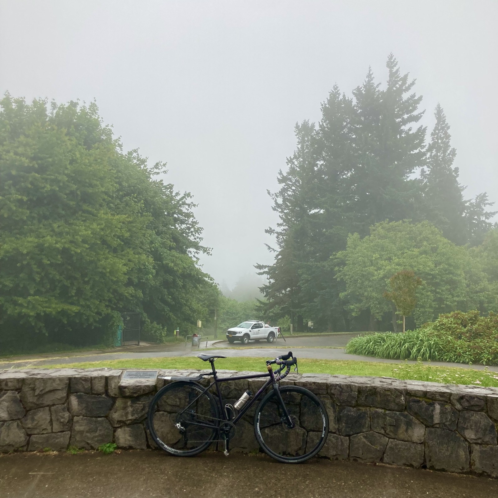 View from Council Crest Park toward Mt. Hood, which is obscured by rain and fog. In the foreground a bicycle leans against a low stone wall. Attached to the bicycle are a brass bell, a small battered steel coffee thermos, and a cheap plastic fender. About 50' distant is a PP&R pickup with gardening equipment. The entire scene is wet and brilliant with lush vegetation.