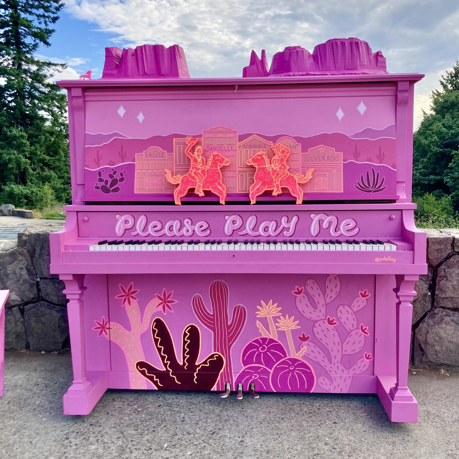 Upright piano outdoors at Council Crest Park, boldly painted with Old West motifs in pink and purple. Cacti, sagebrush, mountains, buttes etc. There are two cowboy cutouts in hot pink and yellow attached above the keyboard. Famous Portland gay bars & clubs are depicted as Old West Saloons: The Eagle, Slaughters, Darcelle, Scandals, Silverado. Attached to the top of the piano is a carved relief of buttes resembling Monument Valley. “Please Play Me” is written in large letters above the keyboard. It is tagged with @pdxflag. (Not in the picture is the artist credit, attached to the side: “This piano was created by Grant Brady”