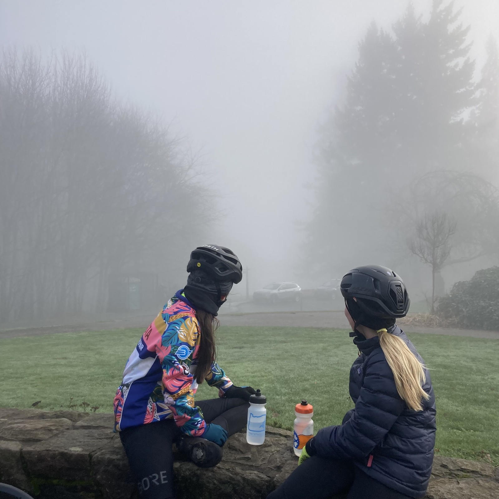 Two young women in warm gear and bicycle helmets seated on a low stone wall, look into the fog toward the direction of Mt. Hood
