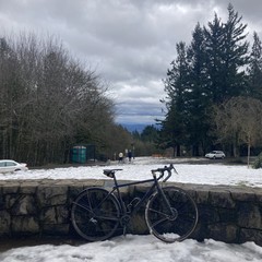 View from Council Crest Park toward Mt. Hood, which is not visible under a layer of stratus clouds. The air is clear, windy, and cold. In the foreground, a bicycle leans against a decorative stone wall. About 50' away two people stand near the portapotty talking. A 10-day-old layer of icy slush coats the hilltop, it is heavily compressed and churned with footprints