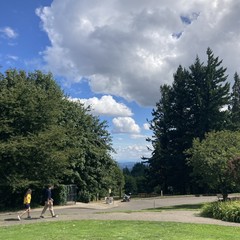 Looking east toward Mt. Hood from Council Crest. Many heavy-but-fluffy wet clouds dot the sky which feels scrubbed clean by the rain of the last two days. In the foreground two people are walking south along a sidewalk
